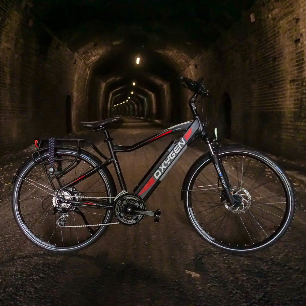 Oxygen S-CROSS CB MKII Trekking Electric Bike, Commuter, Gray 15.5MPH view of the bike from the side on its side stand in a long brick tunnel