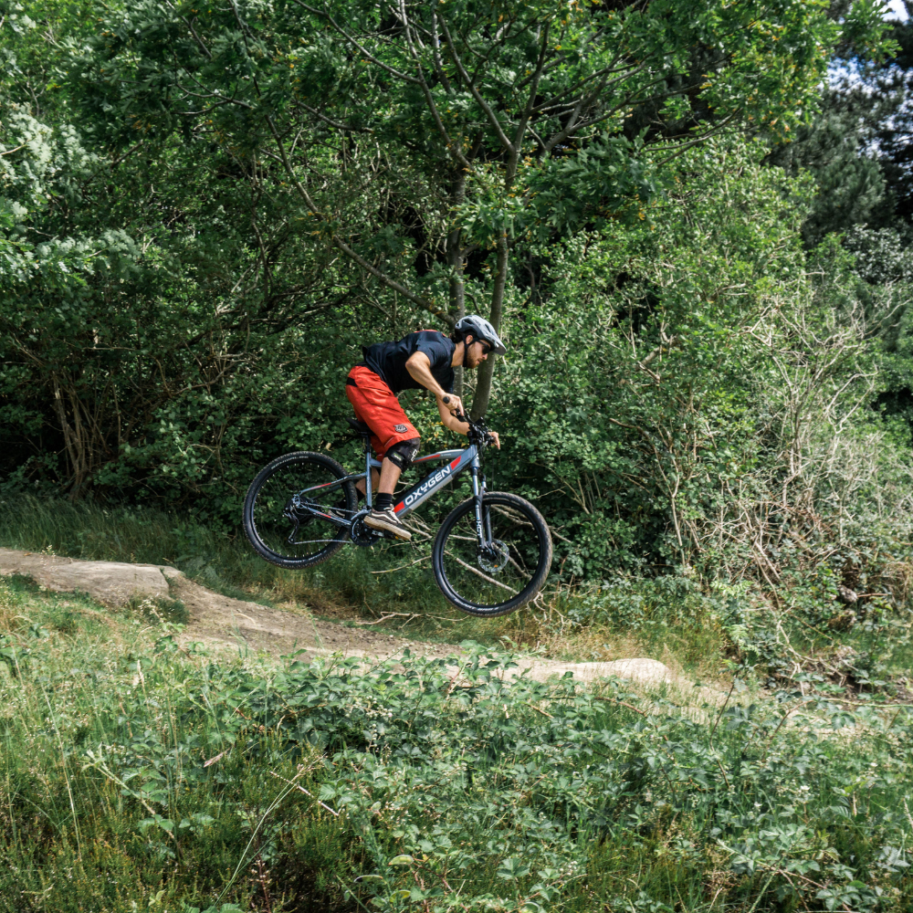 Oxygen S-CROSS MTB MKII All Terrain Electric Bike, Mountain, Gray 15.5MPH Bike and rider jumping off a small hump in a descending woodland trail 