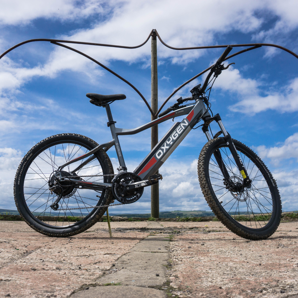 Oxygen S-CROSS MTB MKII All Terrain Electric Bike, Mountain, Gray 15.5MPH View of the bike on a hilltop platform with blue sky behind. Bike facing right on its side stand