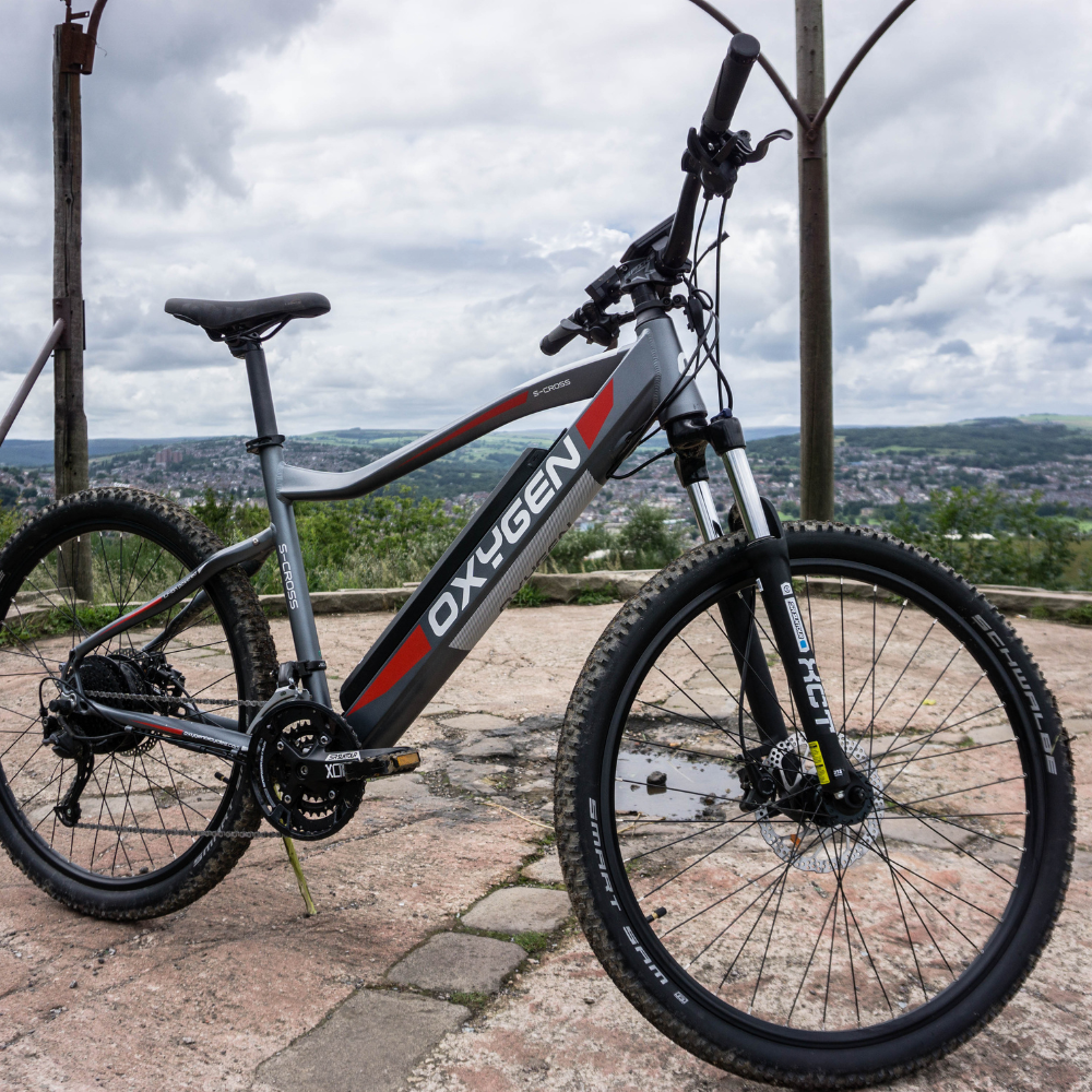 Oxygen S-CROSS MTB MKII All Terrain Electric Bike, Mountain, Gray 15.5MPH Bike on a hilltop platform facing right and overlooking a valley