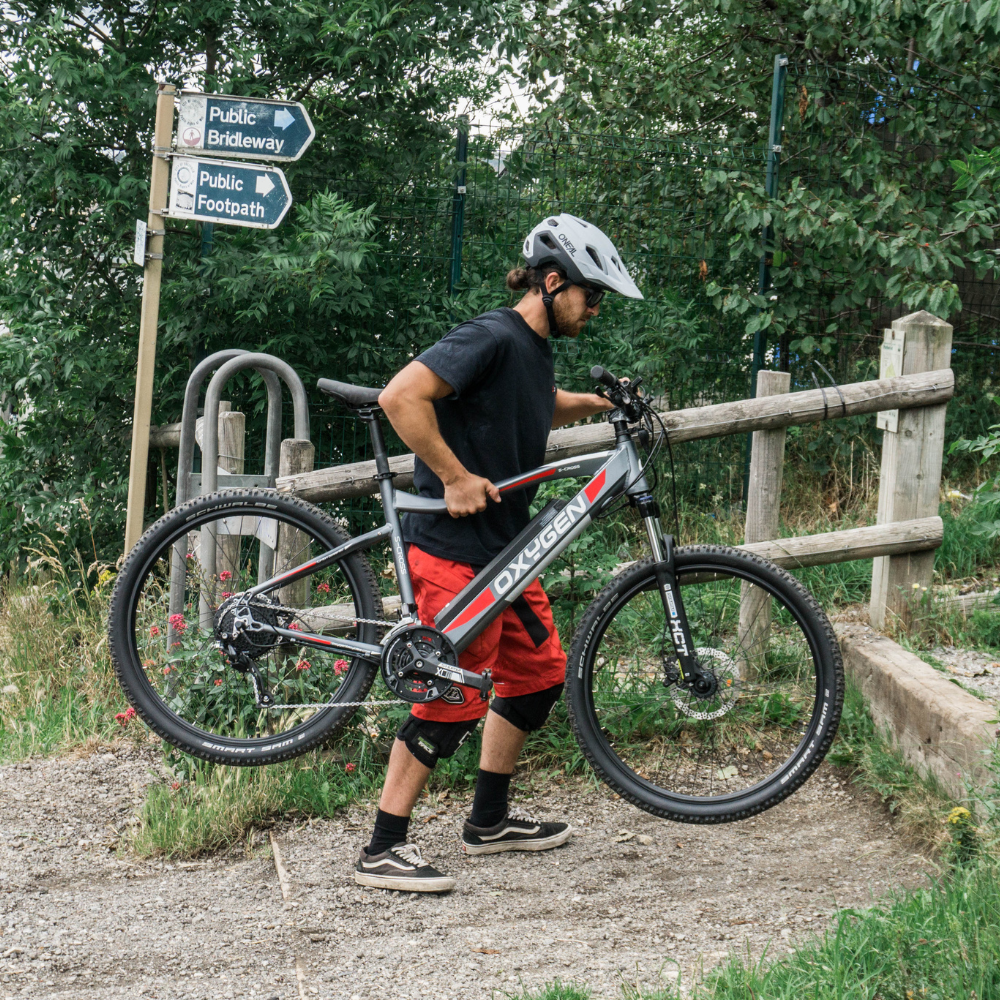 Oxygen S-CROSS MTB MKII All Terrain Electric Bike, Mountain, Gray 15.5MPH Bike is being carried by the rider to maneuver over high style sleeper on their way to a woodland trail