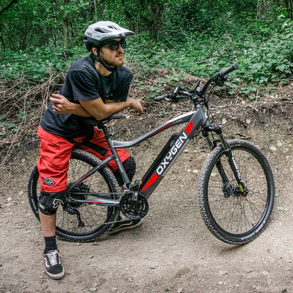 Oxygen S-CROSS MTB MKII All Terrain Electric Bike, Mountain, Gray 15.5MPH Bike with rider on a woodland trail. The rider has stopped and is straddling the rear wheel of the bike and leaning on the saddle