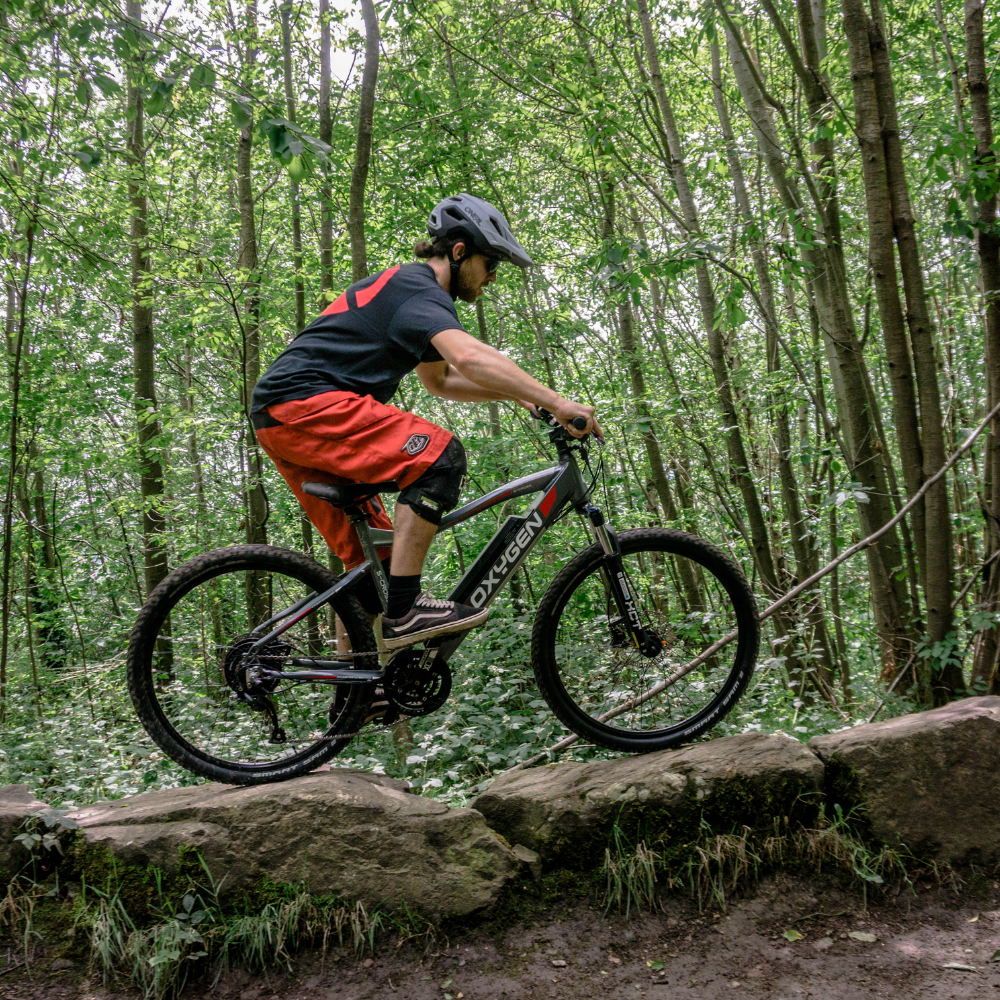 Oxygen S-CROSS MTB MKII All Terrain Electric Bike, Mountain, Gray 15.5MPH Bike is being ridden over some large rocks on a woodland trail ride